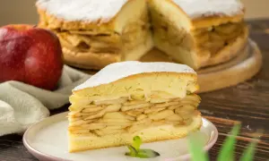 Ein saftiges Stück gedeckter Apfelkuchen mit vielen Apfelschichten, bestäubt mit Puderzucker. Daneben ein roter Apfel.