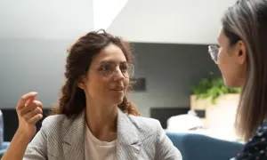 Två kvinnor i samtal, en med glasögon och lockigt hår, den andra med långt hår. En man syns i bakgrunden. Scenen utstrålar autentiskt ledarskap genom öppen kommunikation.