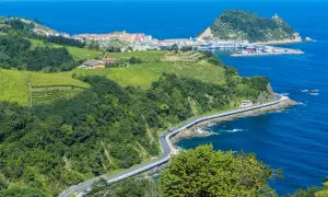 Grüne Weinberge und Wälder säumen die Küstenstraße bei San Sebastián. Das blaue Meer glitzert, während ein kleiner Hafen mit Booten in der Ferne liegt.