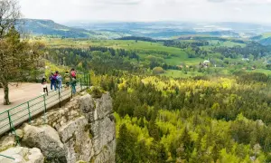 Widok na malownicze szczyty Kotliny Kłodzkiej z platformy widokowej. Grupa turystów podziwia rozległą panoramę lasów i zielonych wzgórz.