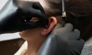 Profesjonalne wykonanie kolczyka orbital piercing. Ręce w czarnych rękawiczkach precyzyjnie przekłuwają ucho.