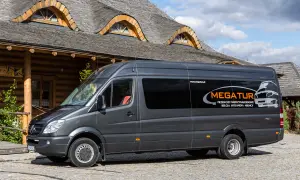 Szary bus firmy MEGATUR, oferujący busy przewozy po polsce i Europie, zaparkowany przed drewnianym budynkiem z charakterystycznym dachem.