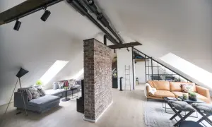 Un salon mansardé avec une cloison de séparation sous pente en briques apparentes, deux canapés, des luminaires industriels et des fenêtres de toit.