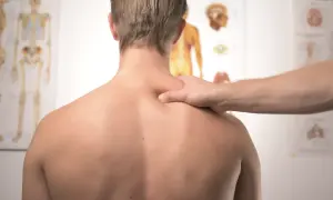 Dłoń terapeuty masuje bark mężczyzny na tle plakatów anatomicznych.