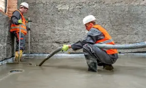 Dwóch budowlańców w kamizelkach odblaskowych wylewa pianobeton. Jeden kontroluje poziom, drugi kieruje wężem.