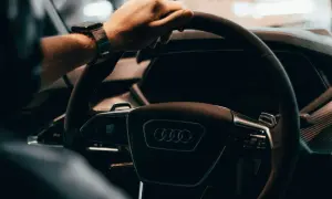 Dłoń na kierownicy Audi, z zegarkiem na nadgarstku. W tle widać fragment deski rozdzielczej, sugerujący wnętrze nowoczesnego samochodu, jak w tabeli z cenami rynkowymi samochodów używanych.