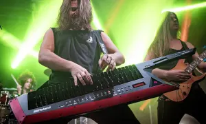Muzyk gra na keyboardzie na pasku, podczas gdy gitarzysta i perkusista tworzą rockową atmosferę.