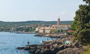 Słoneczna plaża na wyspie Krk, z kamienistym wybrzeżem, turkusowym morzem i miasteczkiem w tle. Wiele osób korzysta z uroków wyspy Krk, zwiedzając jej atrakcje.