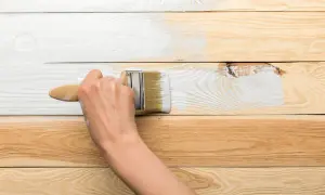 Ręka maluje białą farbą drewnianą boazerię, odnawiając jej wygląd. Proces jak odnowić boazerię krok po kroku.