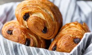 Tre deliziosi pain au chocolat dorati, con strati sfogliati e gocce di cioccolato fondente, adagiati su un canovaccio a righe.