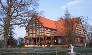 Drewniany dom z czerwonym dachem, otoczony drzewami. To miejsce to jedne z wielu atrakcji Nowego Dworu Gdańskiego.