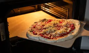 Pizza z sosem i serem na desce, gotowa do włożenia na rozgrzany kamień szamotowy w piekarniku.