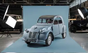En ljusblå Citroën 2CV står i fokus, redo för fotografering. Bakom den syns andra klassiska bilar och studiobelysning.