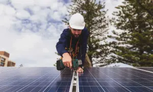 Técnico instala paneles solares, cumpliendo con el real decreto autoconsumo. Edificio y árboles al fondo.