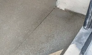 Szara betonowa posadzka z widoczną linią dylatacji, biegnącą od narożnika budynku do metalowej balustrady.