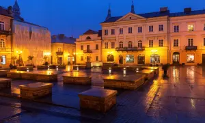 Piotrków Trybunalski atrakcje: Rynek nocą, mokry bruk odbija światła latarni i budynków. Widać kamienne ławki i fasady kamienic.