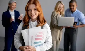 En ung kvinna med rött hår håller i dokument med en vinst- och förlustgraf. Bakom henne diskuterar kollegor affärer, vilket ger tips på svaga sidor.
