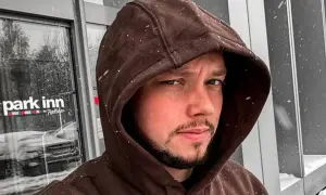 Mężczyzna w brązowej bluzie z kapturem, na tle drzwi hotelu Park Inn. Widać padający śnieg.