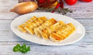 Grillowany ser halloumi na białym talerzu, z dodatkami jak czosnek, cebula, pomidory i chili w tle.