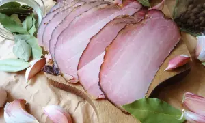 Pokrojony w plastry schab po wędzeniu, otoczony liśćmi laurowymi, czosnkiem i pieprzem.