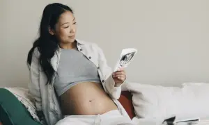 Gravid kvinna tittar på ultraljudsbild, i trimester 2. Hon sitter bekvämt i soffan med sin mage synlig.
