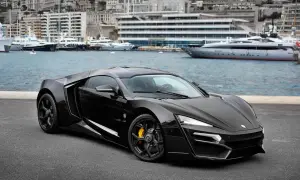 Czarny Lykan Hypersport z żółtymi zaciskami hamulcowymi na tle portu z luksusowymi jachtami i budynkami.