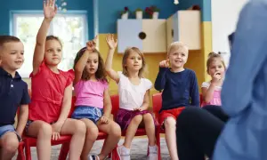 Dzieci w wieku przedszkolnym, które mogą mieć 6 lat, podnoszą ręce, odpowiadając na pytanie nauczyciela.
