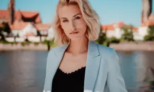 Portret fotograficzny młodej kobiety z blond włosami, w jasnoniebieskiej marynarce, na tle miasta i rzeki.