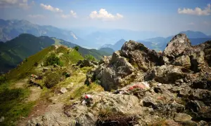 Senderos marcados con rojo y blanco serpentean por las laderas verdes de los Picos de Europa en verano, con rocas imponentes y un valle brumoso al fondo.