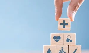 Dłoń układa kostkę z symbolem krzyża na piramidzie z kostek z ikonami zdrowia i bezpieczeństwa. To symboliczne przedstawienie obowiązków pracodawcy w zakresie BHP.