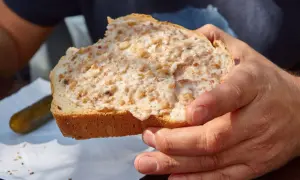 Kromka chleba posmarowana smalcem z kawałkami boczku, trzymana w dłoniach. Obok ogórek kiszony.