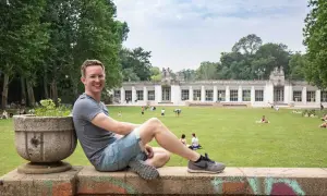 Ein Mann sitzt auf einer Mauer im Park am Stadtbad Charlottenburg Wilmersdorf. Menschen entspannen sich auf der Wiese vor dem Gebäude.