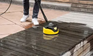 Osoba myje drewniany taras na balkonie w bloku za pomocą żółtej myjki ciśnieniowej.