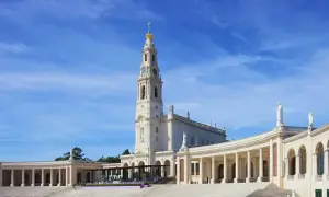 Biała bazylika z wieżą i złotą koroną na szczycie, otoczona kolumnadą. To Sanktuarium Matki Bożej Różańcowej w Fatimie, miejsce pielgrzymek.