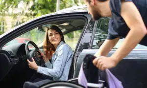 Una donna disabile sorride al volante mentre un uomo aiuta a caricare la sua sedia a rotelle in auto.