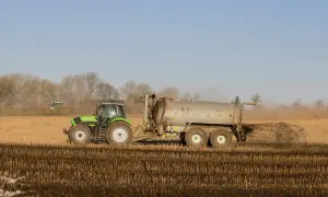 Zielony traktor z przyczepą rozprowadza nawóz na polu. W tle widać drzewa i inne maszyny rolnicze.