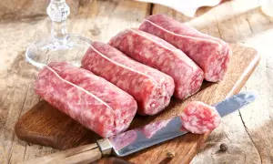Frische Salsiccia-Würste liegen auf einem Holzbrett. Ein Messer mit einem Stück Wurst zeigt, was ist Salsiccia: grob gehacktes Schweinefleisch, gewürzt und in Darm gefüllt.