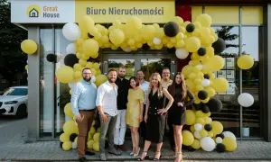 Grupa ludzi przed biurem nieruchomości ozdobionym żółtymi balonami.