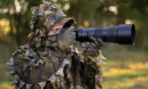Osoba w kamuflażu z liśćmi czeka na ptaki, gotowa do fotografowania.