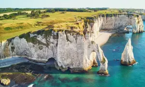 Wapienne klify nad kanałem La Manche, z łukiem skalnym i iglicą. Na szczycie klifów zielone pola golfowe.