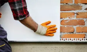 Pracownik w pomarańczowych rękawicach przykłada styropian do ceglanej ściany. Zastanawiasz się, ile kosztuje ankrowanie domu?