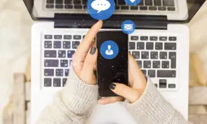 Dłonie trzymające smartfon nad klawiaturą laptopa. Ikony czatu, serca i maila unoszą się w powietrzu. Czy jak usunę czat na Messengerze to druga osoba to widzi?