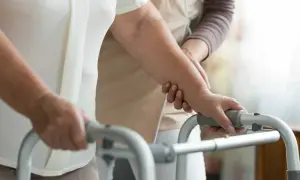 Una persona anziana, con l'aiuto di un'assistente, si appoggia a un deambulatore, mostrando la difficoltà nel riuscire a camminare all'improvviso.