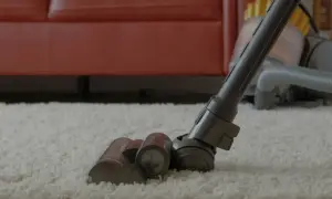 Szara elektroszczotka odkurzacza pracuje na białym dywanie, zbliżając się do czerwonej kanapy.
