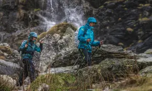 Dwie osoby w turkusowych kurtkach w góry, z kijkami trekkingowymi, idą obok wodospadu.