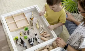Chłopiec z nieleczonym zespołem Aspergera bawi się figurkami zwierząt na piasku, pod okiem terapeuty.