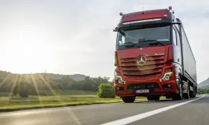 Un camion rouge vif roule sur une route ensoleillée, le soleil couchant illuminant le paysage. Ce camion sans rétroviseur semble prêt pour une longue route.