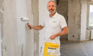 Maler grundiert mit einer Rolle die Wand. Er hält einen Eimer mit "StoPrim GT" in der Hand.