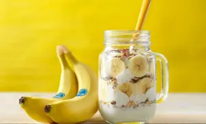 Un delizioso barattolo con yogurt, granola e fette di banana, perfetto per una banana a colazione. Accanto, due banane fresche su sfondo giallo.