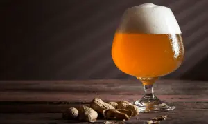 Un bicchiere di keller beer dorata con schiuma bianca, accompagnata da arachidi su un tavolo di legno scuro.